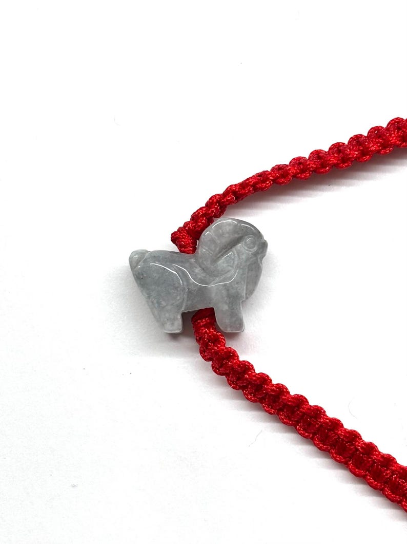May include: A bracelet featuring a bright red woven cord and a single, carved light gray stone charm shaped like a ram. The cord is constructed with a repeating knotted pattern, and the stone charm is positioned centrally on the red string.