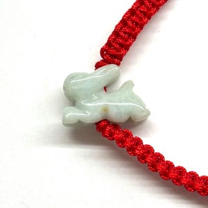 May include: A bright red braided cord bracelet featuring a pale green, translucent carved jade rabbit charm. The rabbit is depicted in a leaping or running pose, with smooth, polished surfaces and subtle natural color variations in the stone.