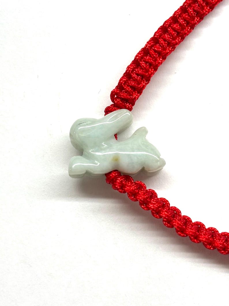 May include: A bright red braided cord bracelet featuring a pale green, translucent carved jade rabbit charm. The rabbit is depicted in a leaping or running pose, with smooth, polished surfaces and subtle natural color variations in the stone.
