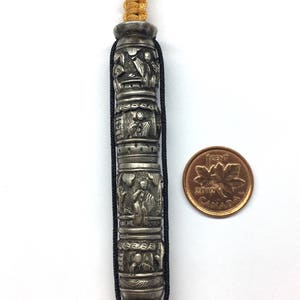May include: A silver charm with intricate carvings of figures and designs. The charm is attached to a black cord with a gold cord accent. A Canadian penny is included for size reference.
