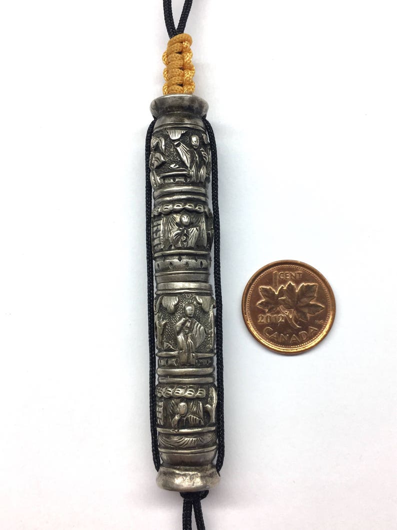 May include: A silver charm with intricate carvings of figures and designs. The charm is attached to a black cord with a gold cord accent. A Canadian penny is included for size reference.