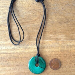 May include: A green malachite stone pendant in the shape of a circle with a hole in the center, hanging from a black cord necklace. A Canadian penny is included for size reference.