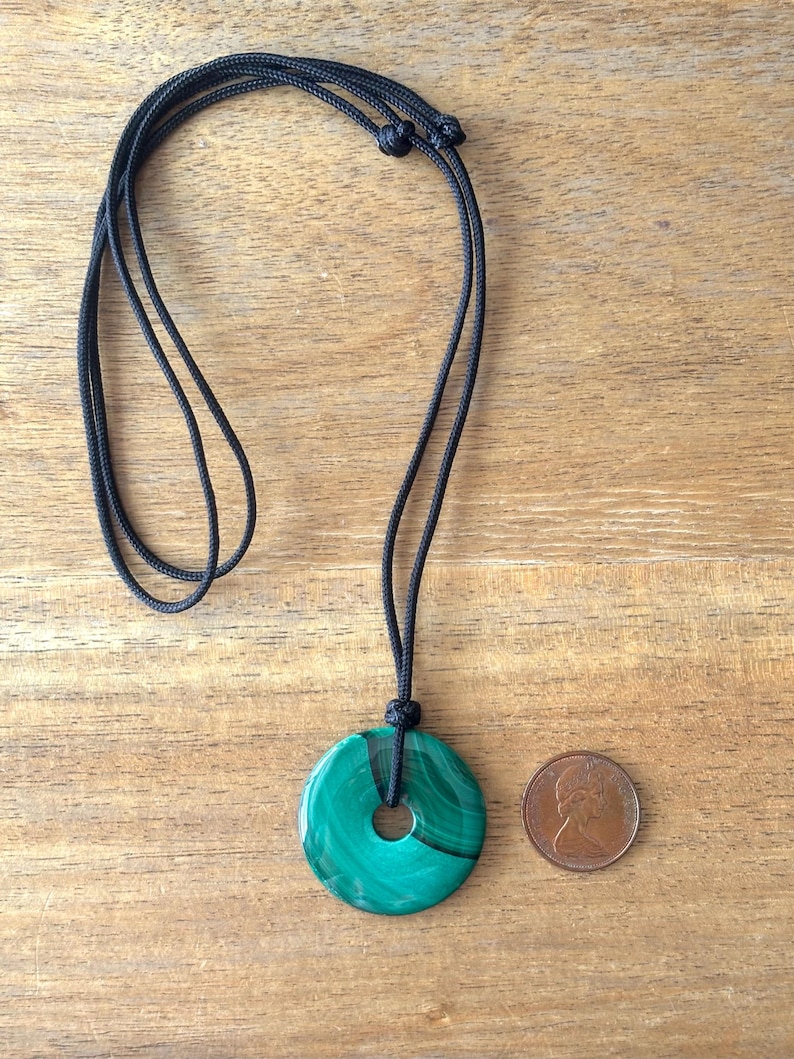 May include: A green malachite stone pendant in the shape of a circle with a hole in the center, hanging from a black cord necklace. A Canadian penny is included for size reference.
