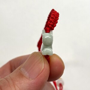 May include: A bracelet featuring a bright red braided cord and a pale green carved stone charm in the shape of a small animal. The charm is centered on the cord, which has a textured, woven appearance throughout its length.