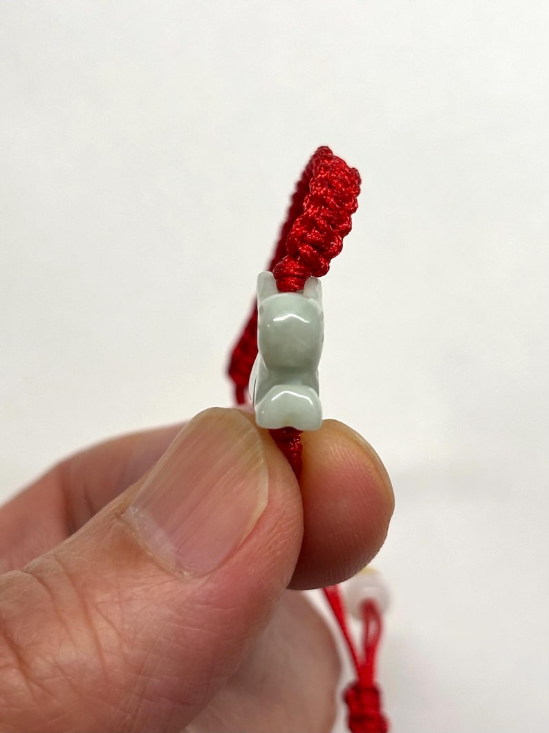May include: A bracelet featuring a bright red braided cord and a pale green carved stone charm in the shape of a small animal. The charm is centered on the cord, which has a textured, woven appearance throughout its length.