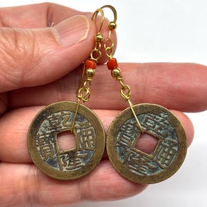 May include: A pair of gold-colored earrings featuring circular, antique-style coins with square cutouts. The earrings have gold-colored hooks and are accented with small red beads. The coins have visible Chinese characters.