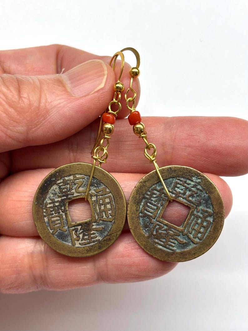 May include: A pair of gold-colored earrings featuring circular, antique-style coins with square cutouts. The earrings have gold-colored hooks and are accented with small red beads. The coins have visible Chinese characters.