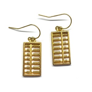 May include: A pair of gold-coloured abacus earrings. Each earring features a rectangular abacus design with small beads and a hook for the ear. The earrings are set against a white background.