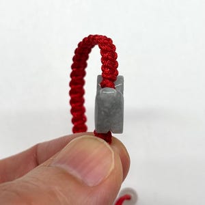 May include: A bracelet featuring a vibrant red braided cord made of nylon thread, accented with a smooth, polished gray stone charm. The stone has a rectangular shape with rounded edges and is threaded directly onto the center of the red cord.