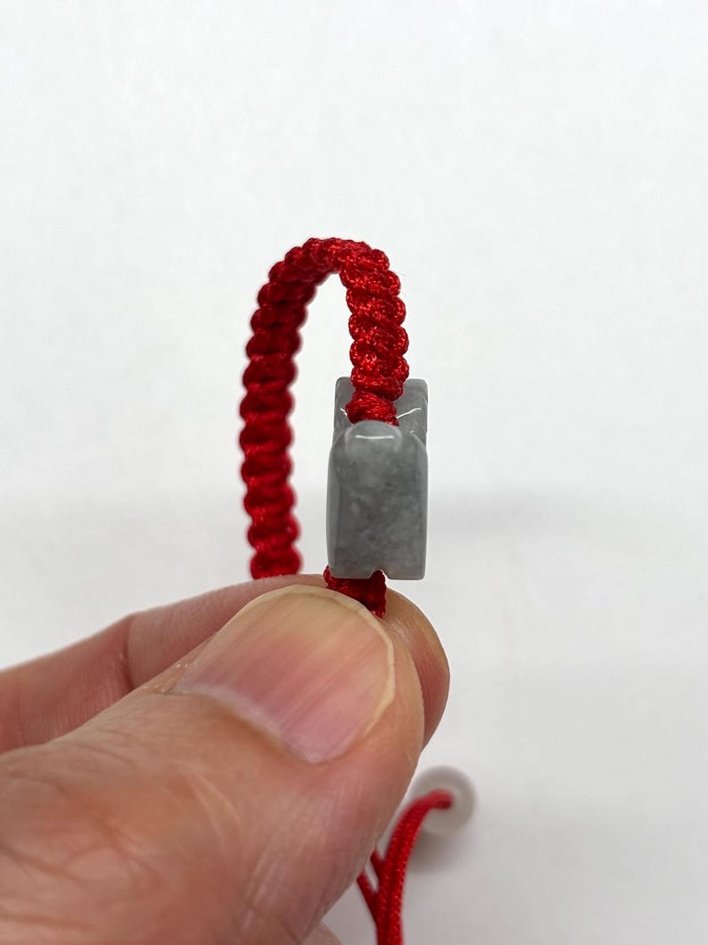 May include: A bracelet featuring a vibrant red braided cord made of nylon thread, accented with a smooth, polished gray stone charm. The stone has a rectangular shape with rounded edges and is threaded directly onto the center of the red cord.