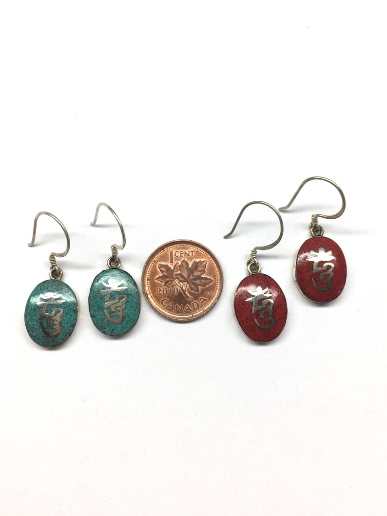 May include: Four silver earrings with oval-shaped charms. Two charms are turquoise blue with silver lettering and two charms are red with silver lettering.