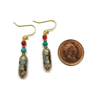 May include: A pair of gold dangle earrings with a white, blue, and red patterned bead and gold beads. The earrings are shown next to a Canadian penny for size reference.