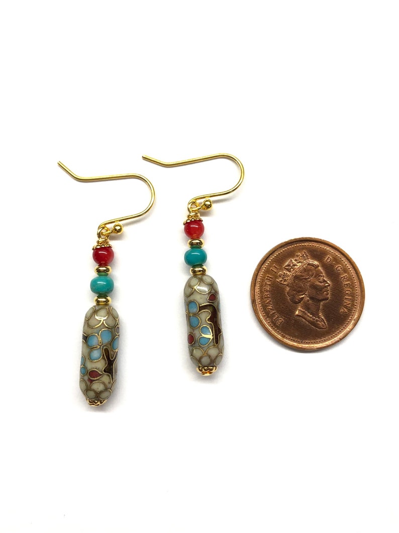 May include: A pair of gold dangle earrings with a white, blue, and red patterned bead and gold beads. The earrings are shown next to a Canadian penny for size reference.