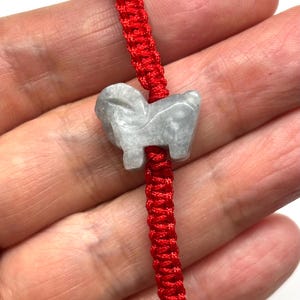 May include: A bright red braided cord bracelet featuring a central light gray stone charm carved into the shape of a ram. The cord has a woven texture and the stone has a smooth, polished finish with subtle variations in its gray color tone.
