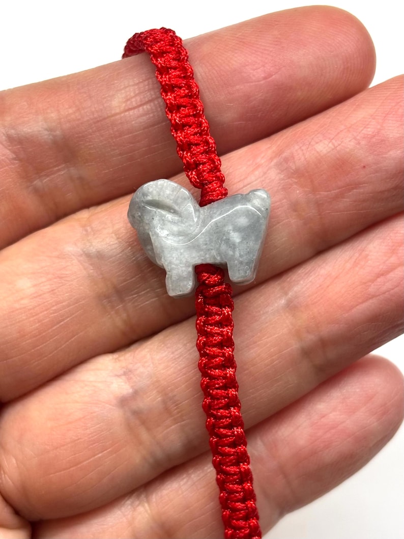 May include: A bright red braided cord bracelet featuring a central light gray stone charm carved into the shape of a ram. The cord has a woven texture and the stone has a smooth, polished finish with subtle variations in its gray color tone.