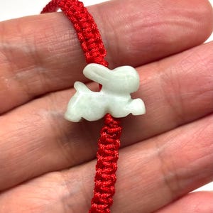 May include: A bracelet featuring a pale green, translucent stone carved into the shape of a rabbit. The rabbit charm is centered on a vibrant red cord that is woven with a repeating knot pattern throughout the length of the band.