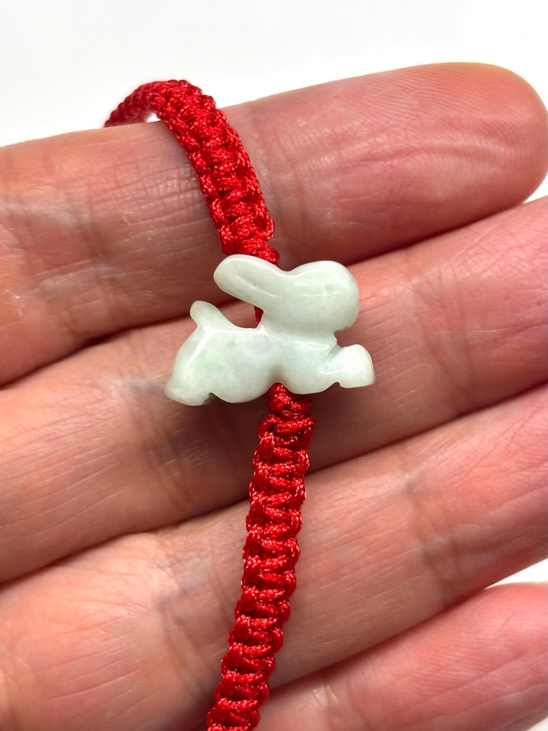 May include: A bracelet featuring a pale green, translucent stone carved into the shape of a rabbit. The rabbit charm is centered on a vibrant red cord that is woven with a repeating knot pattern throughout the length of the band.