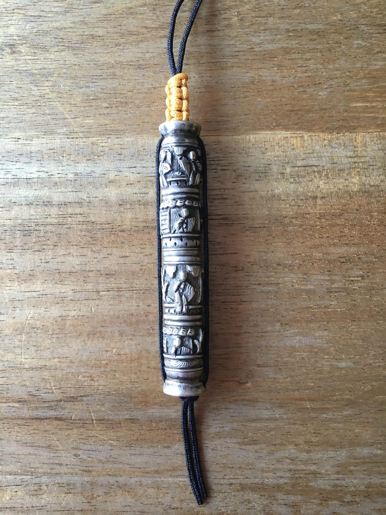 May include: A silver prayer wheel with intricate carvings and a black cord with yellow accents.