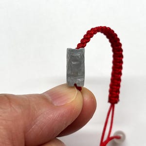 May include: A bracelet featuring a small, rectangular, light gray stone bead carved with a stylized animal figure. The bead is attached to a vibrant red braided cord, which includes an adjustable sliding knot mechanism for a secure fit.