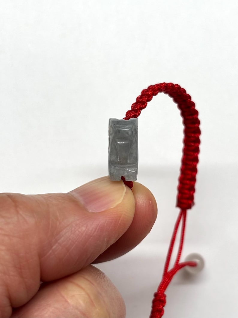 May include: A bracelet featuring a small, rectangular, light gray stone bead carved with a stylized animal figure. The bead is attached to a vibrant red braided cord, which includes an adjustable sliding knot mechanism for a secure fit.