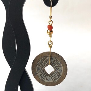 May include: A single gold-colored earring featuring a vintage coin pendant. The earring has a gold wire hook, with red and gold beads. The coin pendant is round with a square hole in the center and Chinese characters.