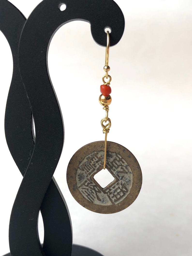 May include: A single gold-colored earring featuring a vintage coin pendant. The earring has a gold wire hook, with red and gold beads. The coin pendant is round with a square hole in the center and Chinese characters.
