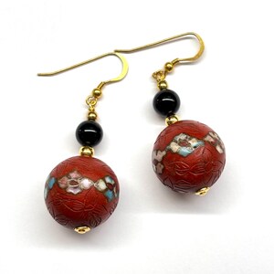 May include: A pair of dangle earrings with red and black beads. The red beads are decorated with floral patterns and white enamel accents. The earrings have gold-toned hooks.
