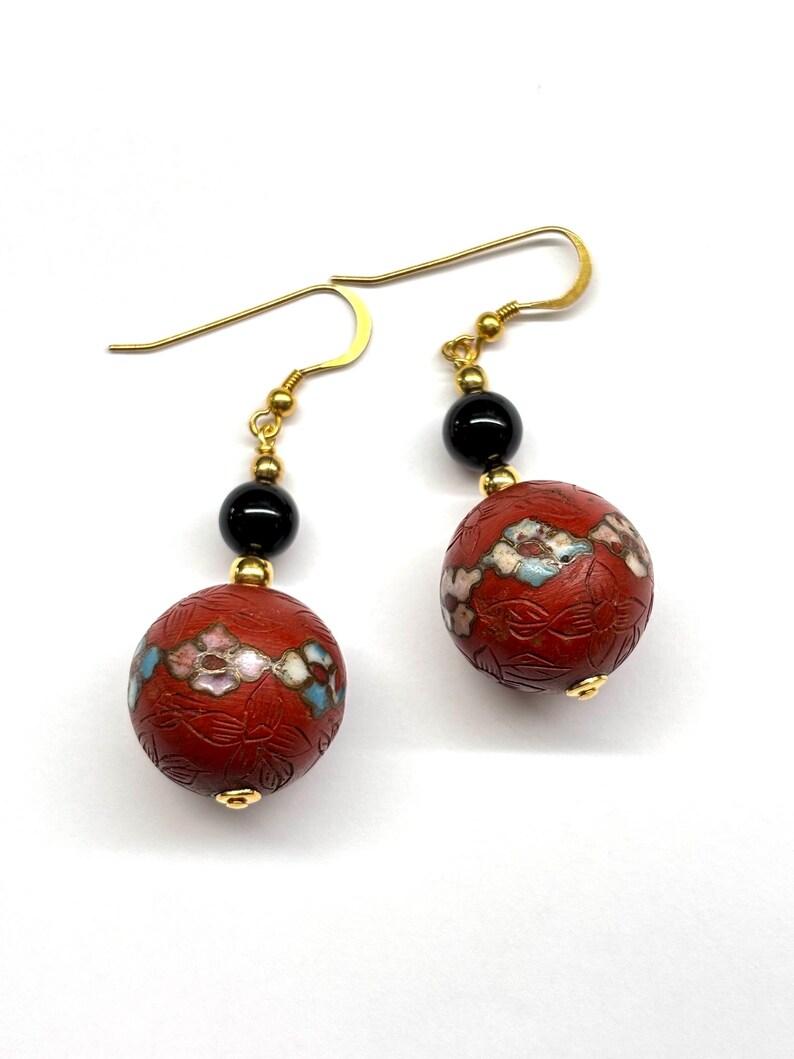 May include: A pair of dangle earrings with red and black beads. The red beads are decorated with floral patterns and white enamel accents. The earrings have gold-toned hooks.