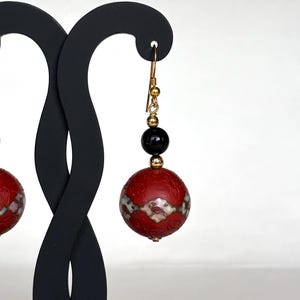 May include: A pair of red and black dangle earrings with a gold hook and a black bead. The red bead is decorated with a white and gold floral pattern.
