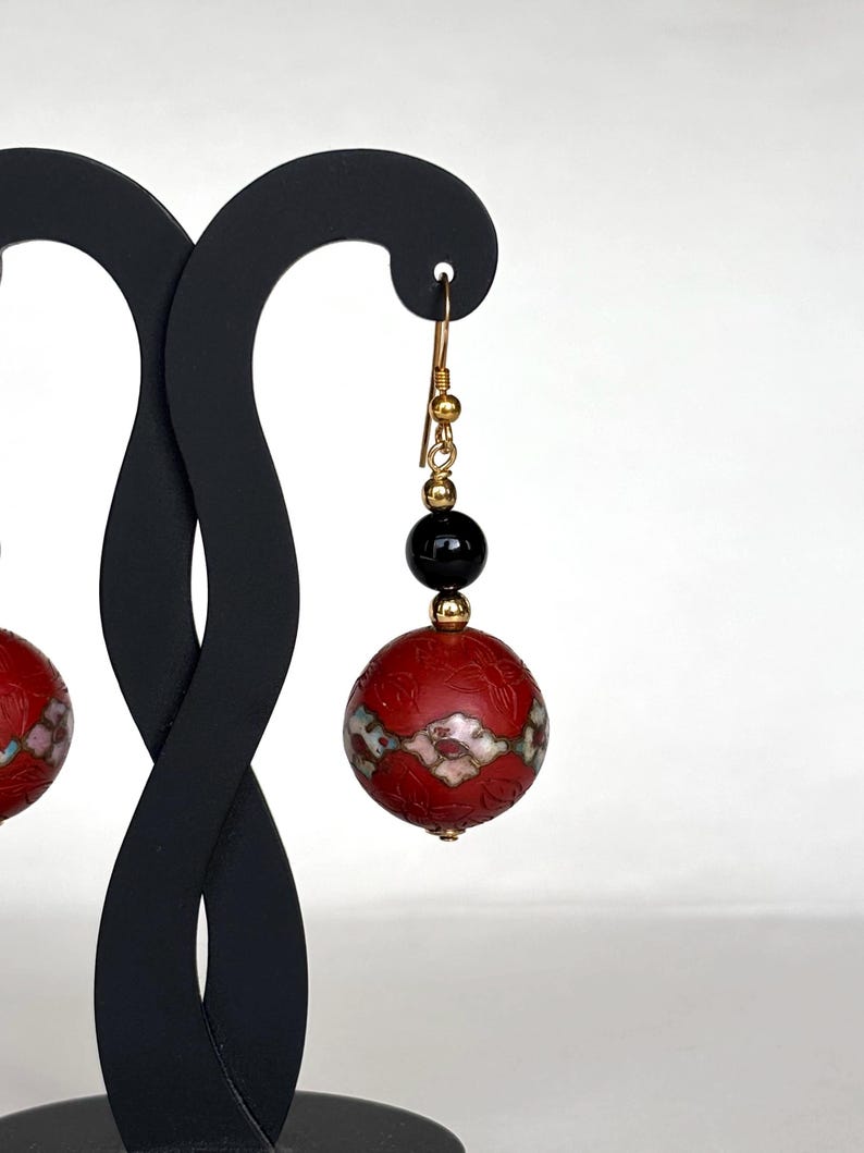 May include: A pair of red and black dangle earrings with a gold hook and a black bead. The red bead is decorated with a white and gold floral pattern.