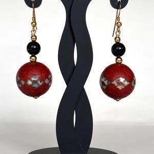 May include: A pair of red and black dangle earrings with gold accents. The earrings feature a round red bead with a white and blue geometric pattern and a black bead above it.