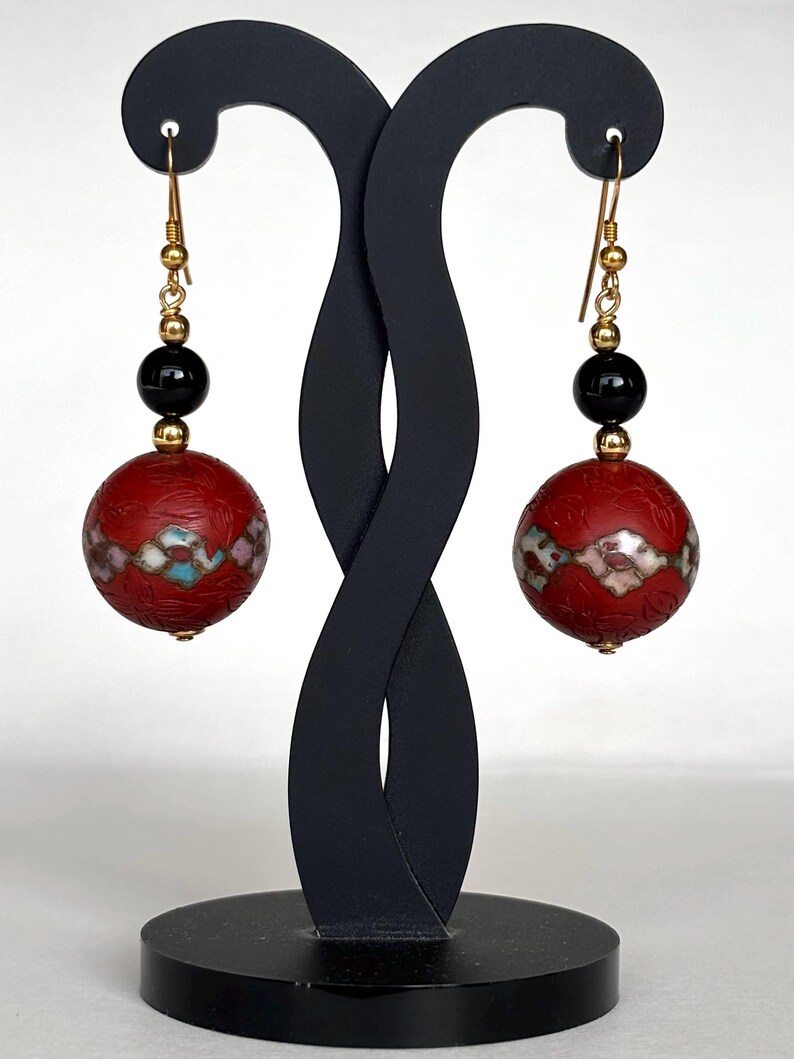 May include: A pair of red and black dangle earrings with gold accents. The earrings feature a round red bead with a white and blue geometric pattern and a black bead above it.