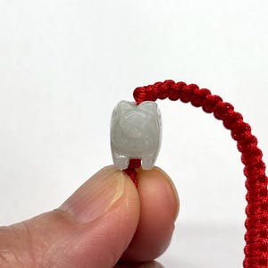 May include: A small, translucent, pale grey carved stone bead shaped like a pig, threaded onto a vibrant red braided cord. The pig-shaped charm features subtle carved details and is positioned centrally on the bright red woven string.