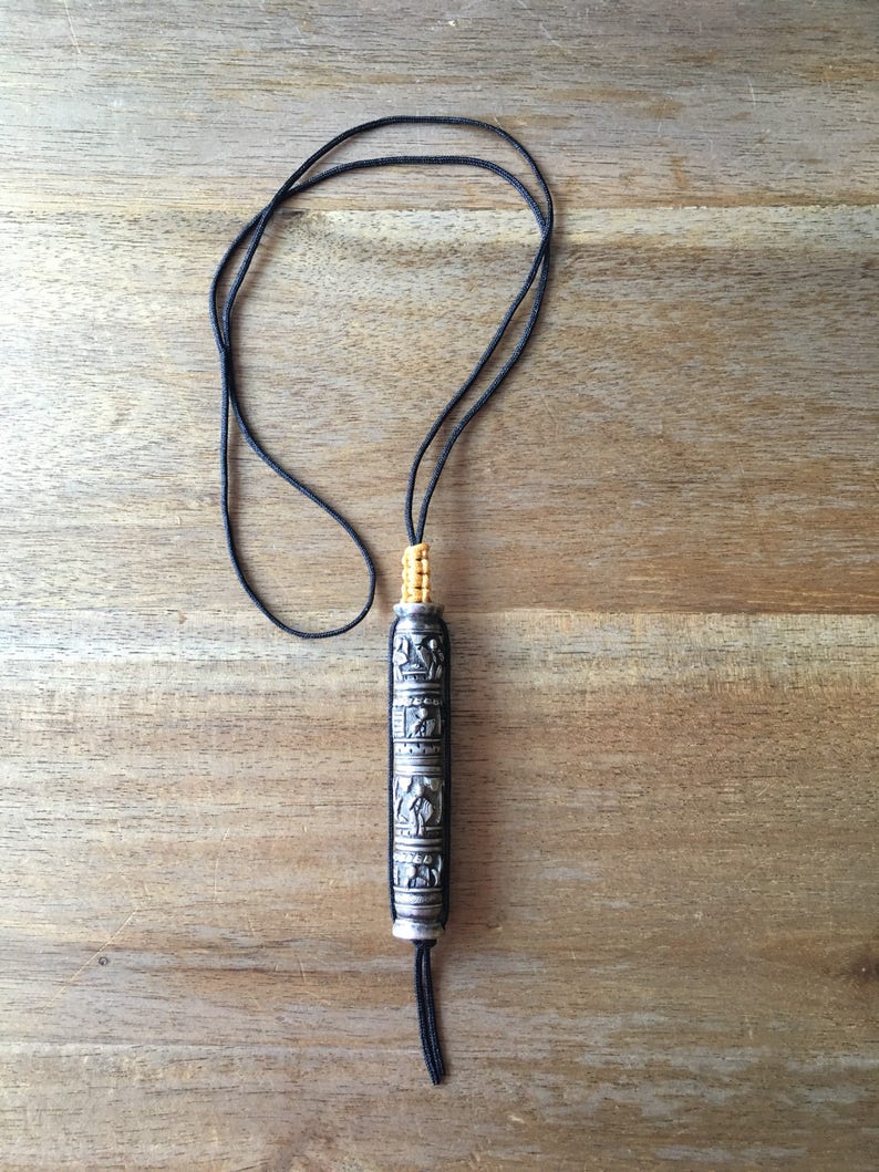 May include: A silver prayer wheel with intricate carvings and a black cord. The prayer wheel is suspended from a black cord and has a yellow tassel.