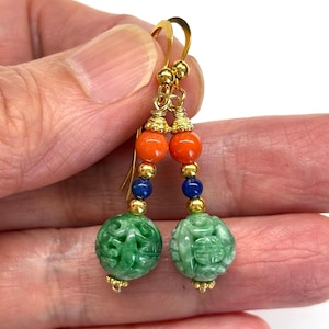 May include: A pair of drop earrings featuring gold-tone hooks and hardware. Each earring consists of a round orange bead, a small blue bead, and a larger, spherical green bead with intricate carved patterns, separated by small gold-tone spacer beads.