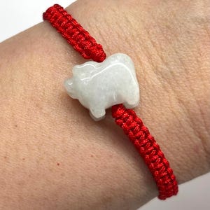 May include: A bracelet featuring a bright red woven cord and a central white stone charm carved into the shape of a small pig. The cord is constructed with a series of tight, uniform knots that secure the stone in place on the wrist.