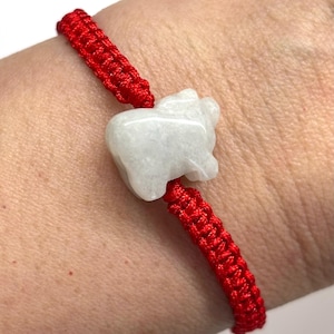May include: A bracelet featuring a bright red woven cord and a central white stone charm carved into the shape of a small animal. The cord is constructed with a series of tight, interlocking knots that secure the stone in place on the wrist.