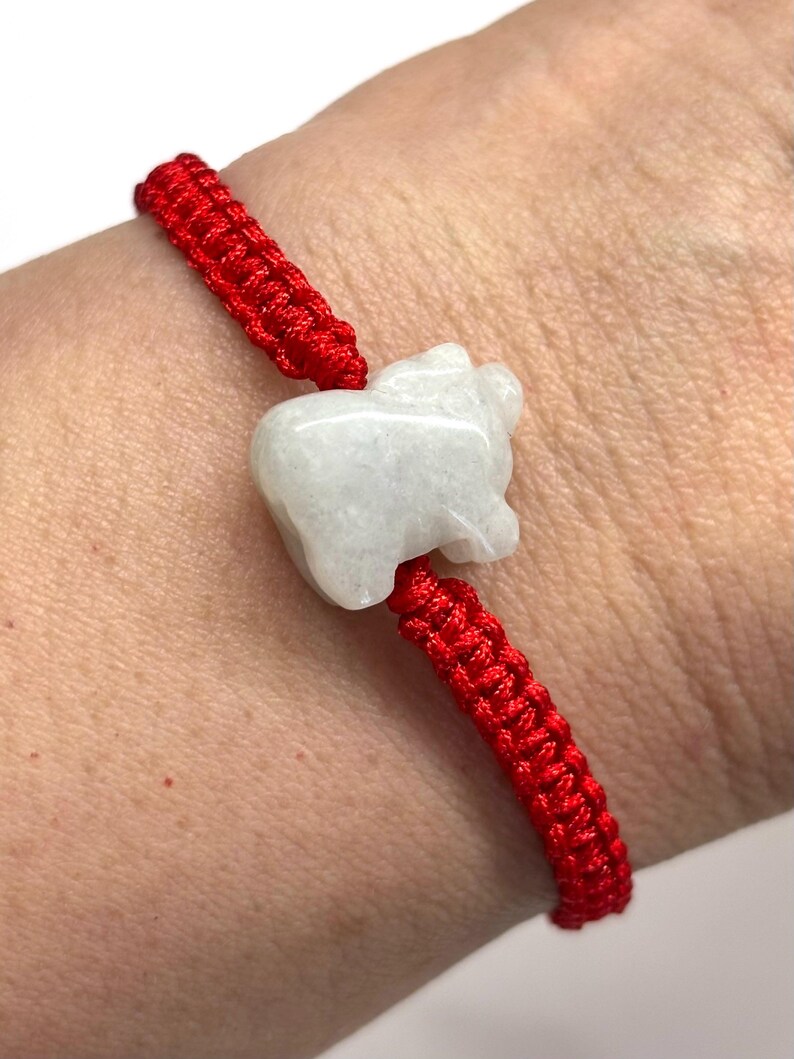 May include: A bracelet featuring a bright red woven cord and a central white stone charm carved into the shape of a small animal. The cord is constructed with a series of tight, interlocking knots that secure the stone in place on the wrist.