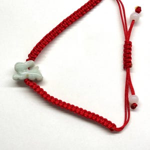 May include: A red cord bracelet featuring a pale green carved rabbit charm. The bracelet is constructed with a braided knot pattern and includes an adjustable sliding closure with two small, round, light-colored beads at the ends of the cords.