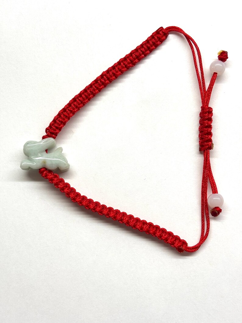 May include: A red cord bracelet featuring a pale green carved rabbit charm. The bracelet is constructed with a braided knot pattern and includes an adjustable sliding closure with two small, round, light-colored beads at the ends of the cords.