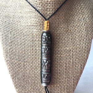 May include: A silver necklace with a cylindrical pendant featuring intricate carvings. The pendant is suspended from a black cord with yellow beads at the top.