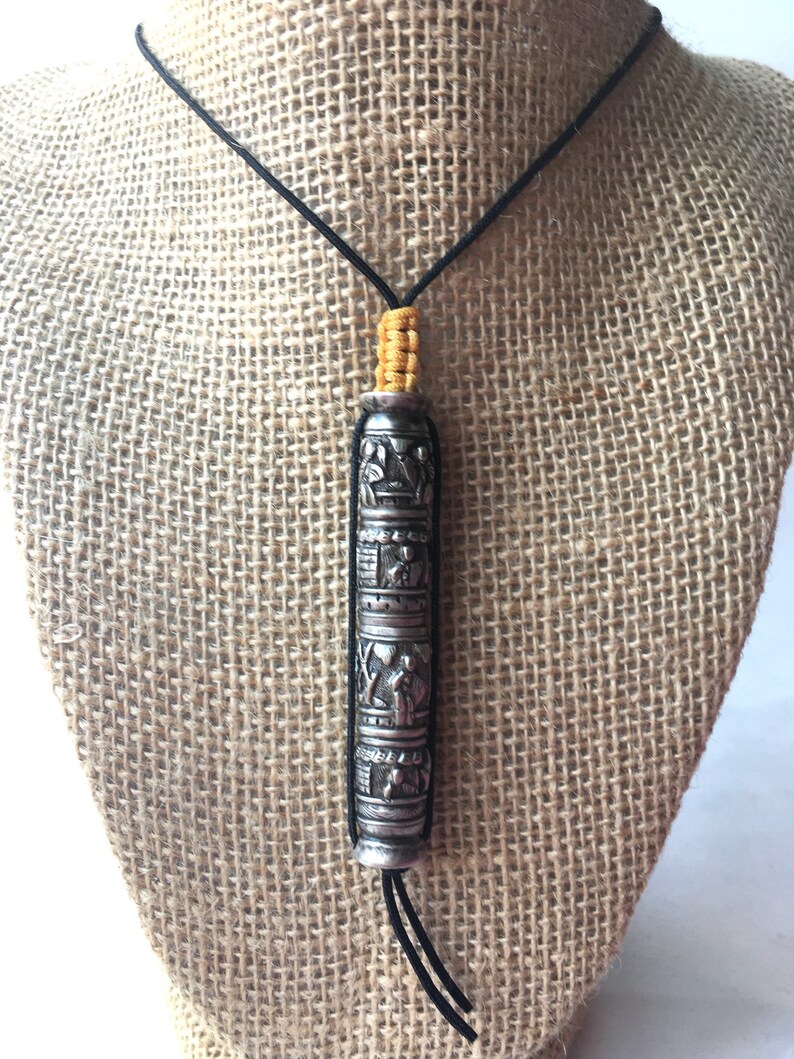 May include: A silver necklace with a cylindrical pendant featuring intricate carvings. The pendant is suspended from a black cord with yellow beads at the top.