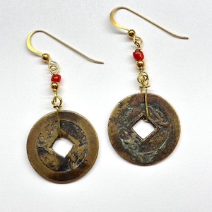May include: A pair of antique-style earrings. Each earring features a circular, bronze-colored coin pendant with a square center hole. Small red beads and gold-colored hooks complete the design. The earrings are displayed against a white background.