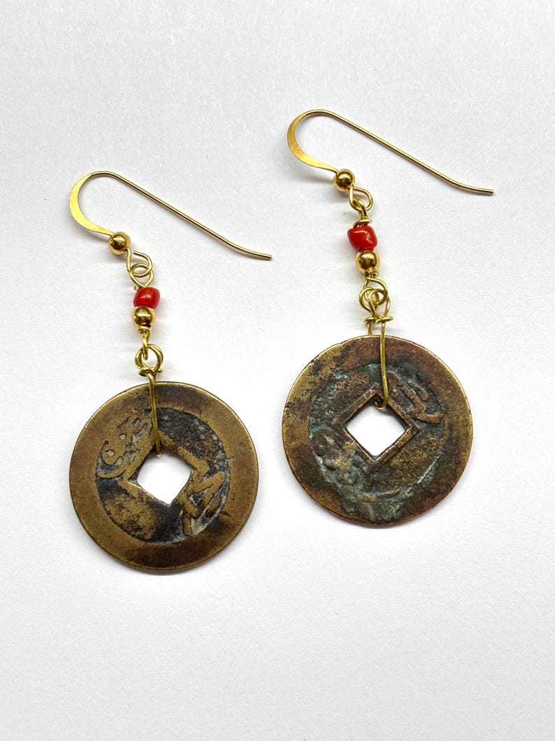 May include: A pair of antique-style earrings. Each earring features a circular, bronze-colored coin pendant with a square center hole. Small red beads and gold-colored hooks complete the design. The earrings are displayed against a white background.