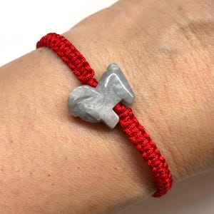 May include: A bright red braided cord bracelet featuring a central light gray stone charm carved into the shape of a ram. The cord is woven in a tight, repeating pattern, and the stone has a smooth, polished finish with subtle gray marbling.