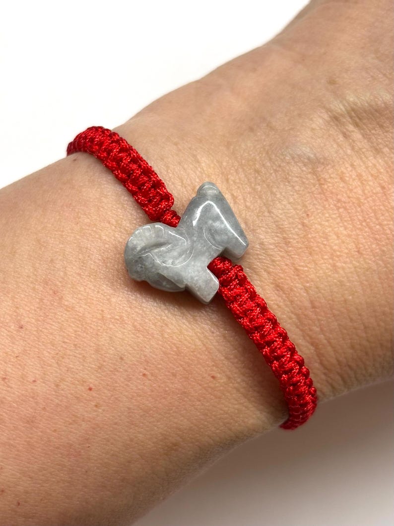 May include: A bright red braided cord bracelet featuring a central light gray stone charm carved into the shape of a ram. The cord is woven in a tight, repeating pattern, and the stone has a smooth, polished finish with subtle gray marbling.