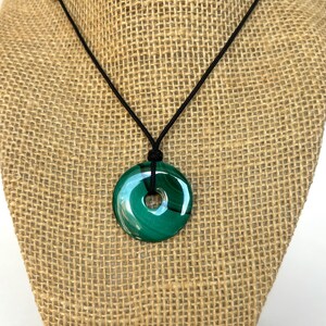 May include: A green malachite stone pendant necklace with a black cord. The pendant is a round, flat disc with a hole in the center.