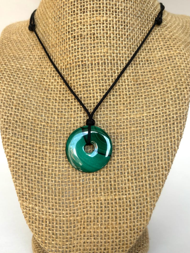 May include: A green malachite stone pendant necklace with a black cord. The pendant is a round, flat disc with a hole in the center.