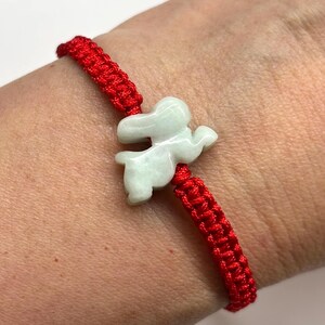 May include: A bracelet featuring a bright red woven cord and a pale green carved stone charm in the shape of a rabbit. The charm is centered on the cord, which has a braided texture. The piece is designed to be worn around the wrist.