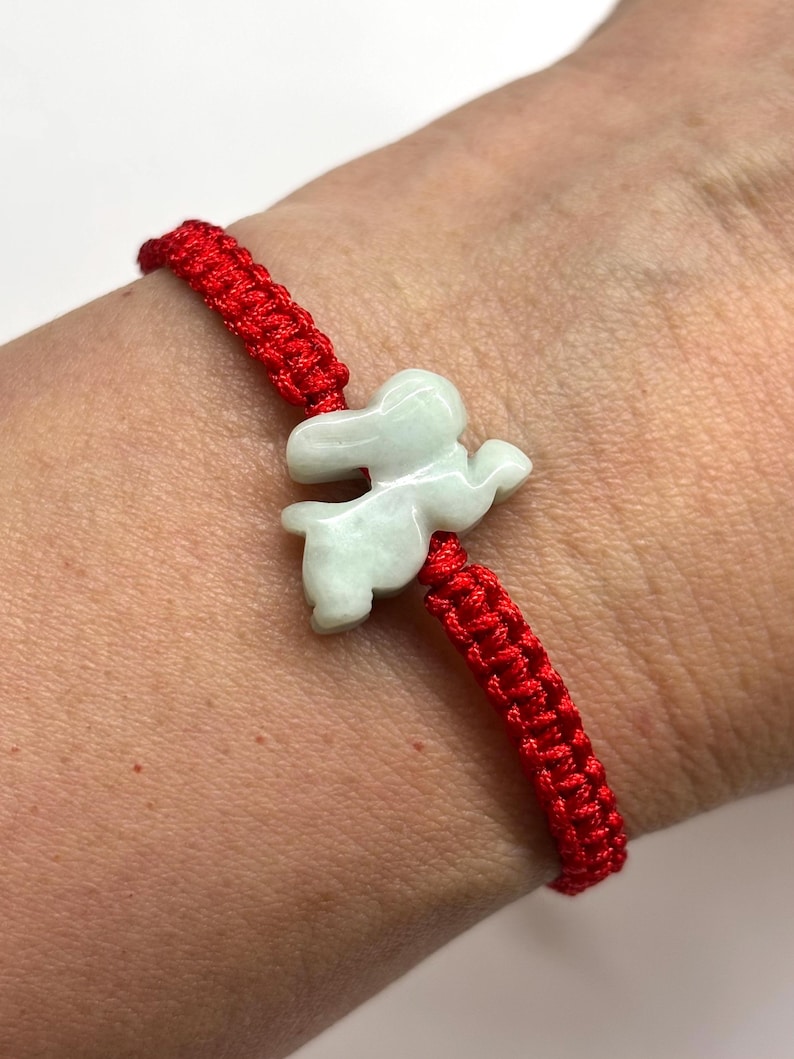 May include: A bracelet featuring a bright red woven cord and a pale green carved stone charm in the shape of a rabbit. The charm is centered on the cord, which has a braided texture. The piece is designed to be worn around the wrist.